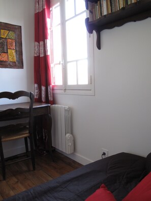 A room with a wooden desk, a chair, a radiator, a bed with red pillows, and a bookshelf with books.