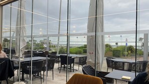 Een terras met tafels en stoelen, grote ramen en uitzicht op zee.