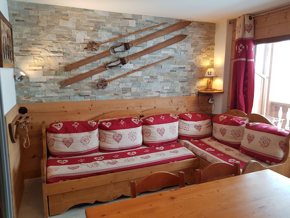 A wooden bench with heart-patterned cushions, a stone wall with wooden skis, and a window with red curtains.