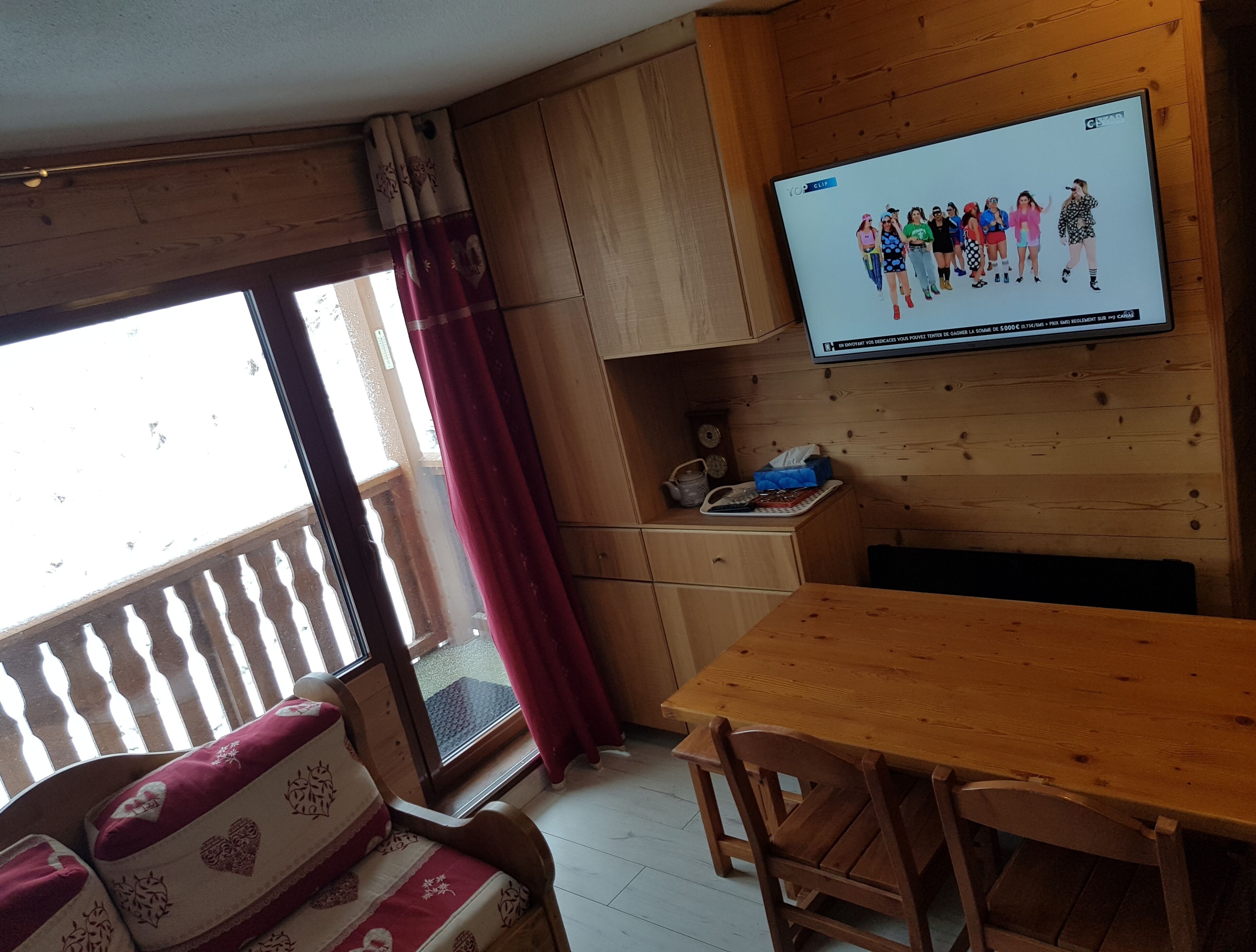 Un intérieur en bois avec un téléviseur fixé au mur, diffusant une image d’un groupe de personnes. On y trouve une table en bois et un banc rembourré orné de motifs en forme de cœur.