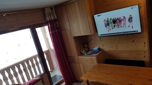 A wooden interior with a TV mounted on the wall displaying a group of people. There is a wooden table and a cushioned bench with heart patterns. - Les menuires (Les Belleville)