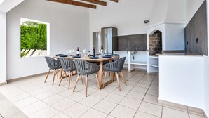Ein moderner Außenbereich zum Essen mit einem Holztisch und Stühlen, einem eingebauten Grill und einem großen Fenster mit Blick auf Grünflächen.