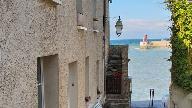 Une ruelle pavée, avec un bâtiment sur la gauche, un banc et une vue sur la mer, où l’on aperçoit un phare au loin.