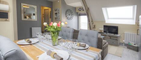 A dining area with a wooden table set for six, grey upholstered chairs, a vase with flowers, and a clock on the wall.
