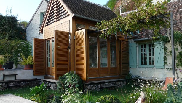 Une maison en bois dotée de grandes fenêtres et de volets, entourée d’une végétation luxuriante et agrémentée d’une terrasse en bois.