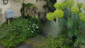 Un bâtiment de style rustique, doté d’une façade en pierre, d’un petit jardin agrémenté de plantes fleuries et d’un élément décoratif aquatique bien visible.