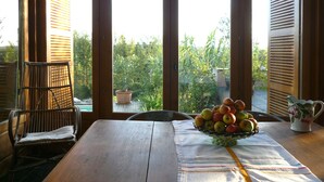 Une table en bois sur laquelle se trouve un saladier rempli de fruits, une chaise en osier et, par de grandes fenêtres, une vue sur la verdure.