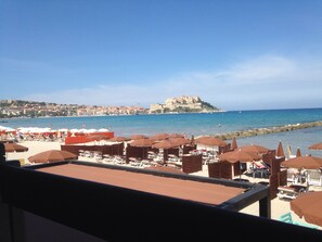 Una playa con sombrillas y sillas de descanso, vista al mar y un castillo en la colina al fondo.