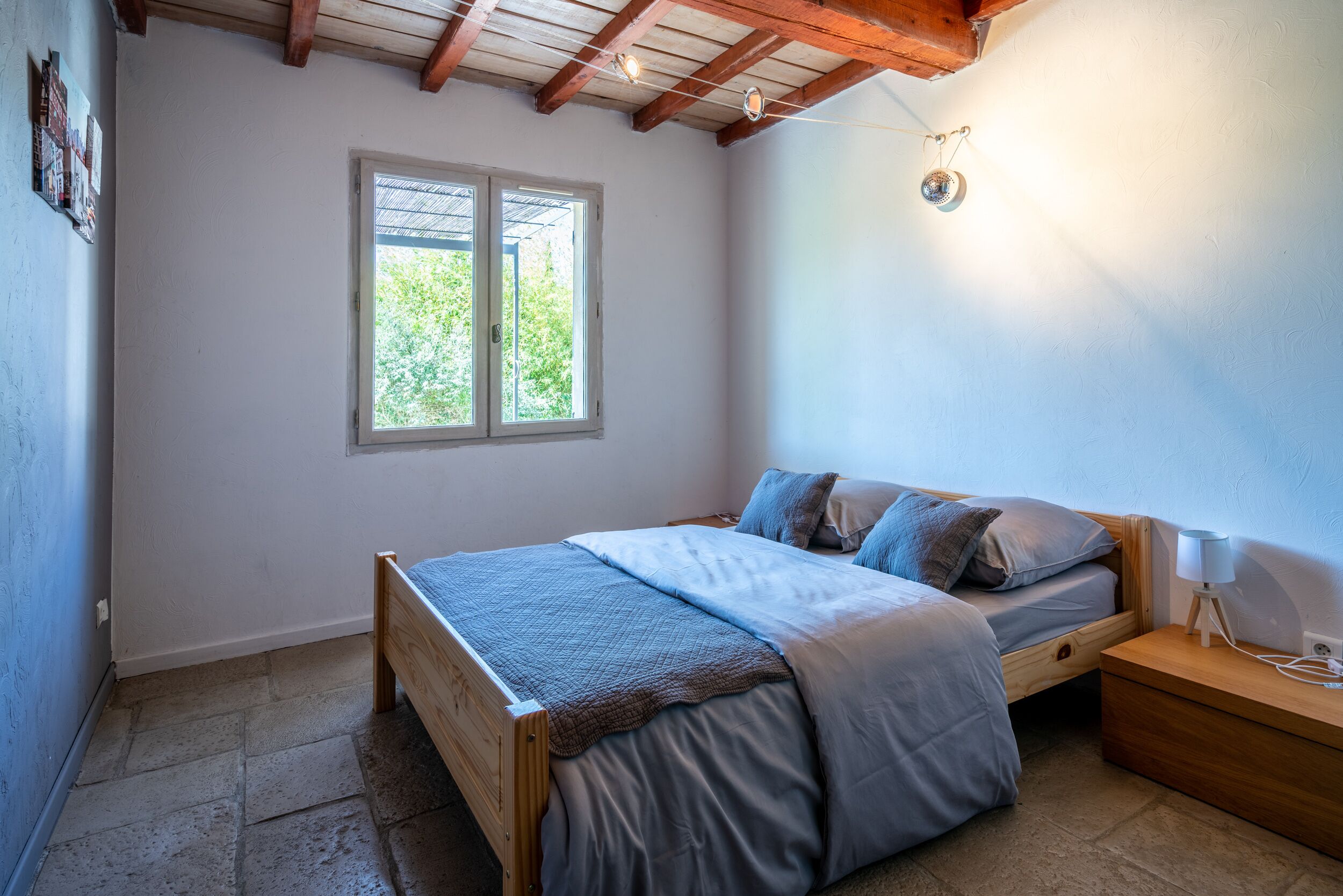 A bedroom with a wooden bed, a nightstand, a lamp, and a window with a view of greenery.