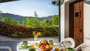 Ein Frühstückstisch mit Obst, Kaffee und Saft, mit Blick auf einen Garten mit Blumen und Hügeln.