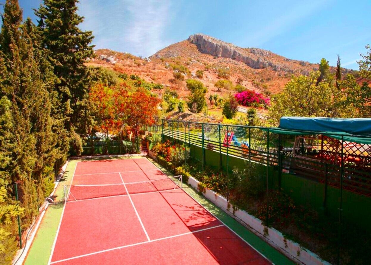 Una cancha de tenis con superficie roja, rodeada por una cerca verde y árboles.