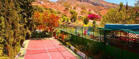 Una cancha de tenis con superficie roja, rodeada por una cerca verde y árboles.