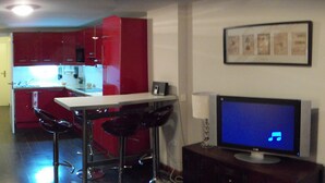 Cocina con armarios rojos, encimera blanca y barra con taburetes. Se ve un televisor sobre un mueble a la derecha.