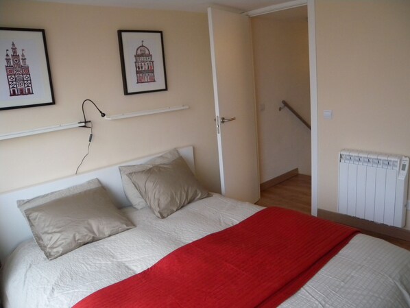 A bedroom with a bed, pillows, a red blanket, a white radiator, and two framed pictures on the wall.