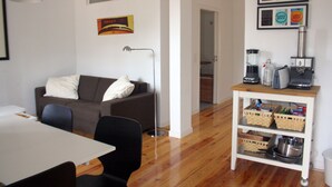 A living room with a sofa, a coffee table, and a dining table with chairs. There is a kitchenette with a coffee maker, a kettle, and a toaster. The room has wooden flooring and white walls. - Old Town of Lisbon, stunning 180º view of Castelo, 3-room apartment with terrace (Lisbonne)