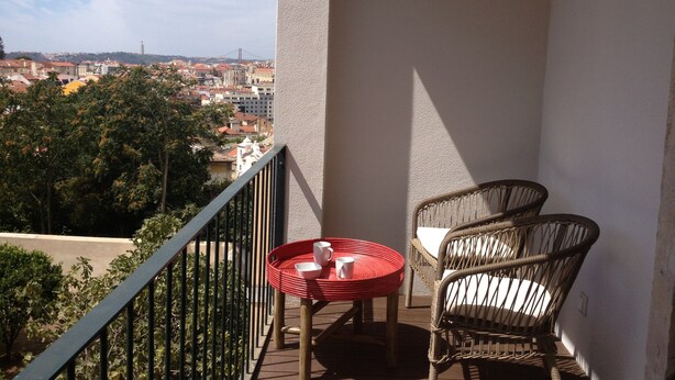 A balcony with a wicker chair and a small table with a red tray, overlooking a cityscape with buildings and trees. - Old Town of Lisbon, stunning 180º view of Castelo, 3-room apartment with terrace (Lisbonne)
