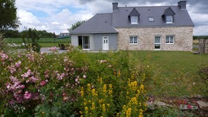 A two-story stone house with a grey roof, a small attached garage, and a garden with pink and yellow flowers.