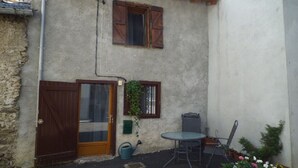 Ein rustikales Gebäude mit Holztür, Fenster mit Fensterläden, einem kleinen Tisch und Stuhl sowie Topfpflanzen.