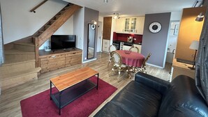 Ein Wohnzimmer mit Holzboden, einer Holztreppe, einem Fernseher, einem Esstisch mit Stühlen, einer Couch und einer Küche im Hintergrund.