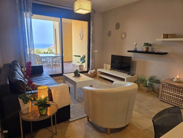 A modern living room with a sofa, a white armchair, a TV, and a balcony with a view.