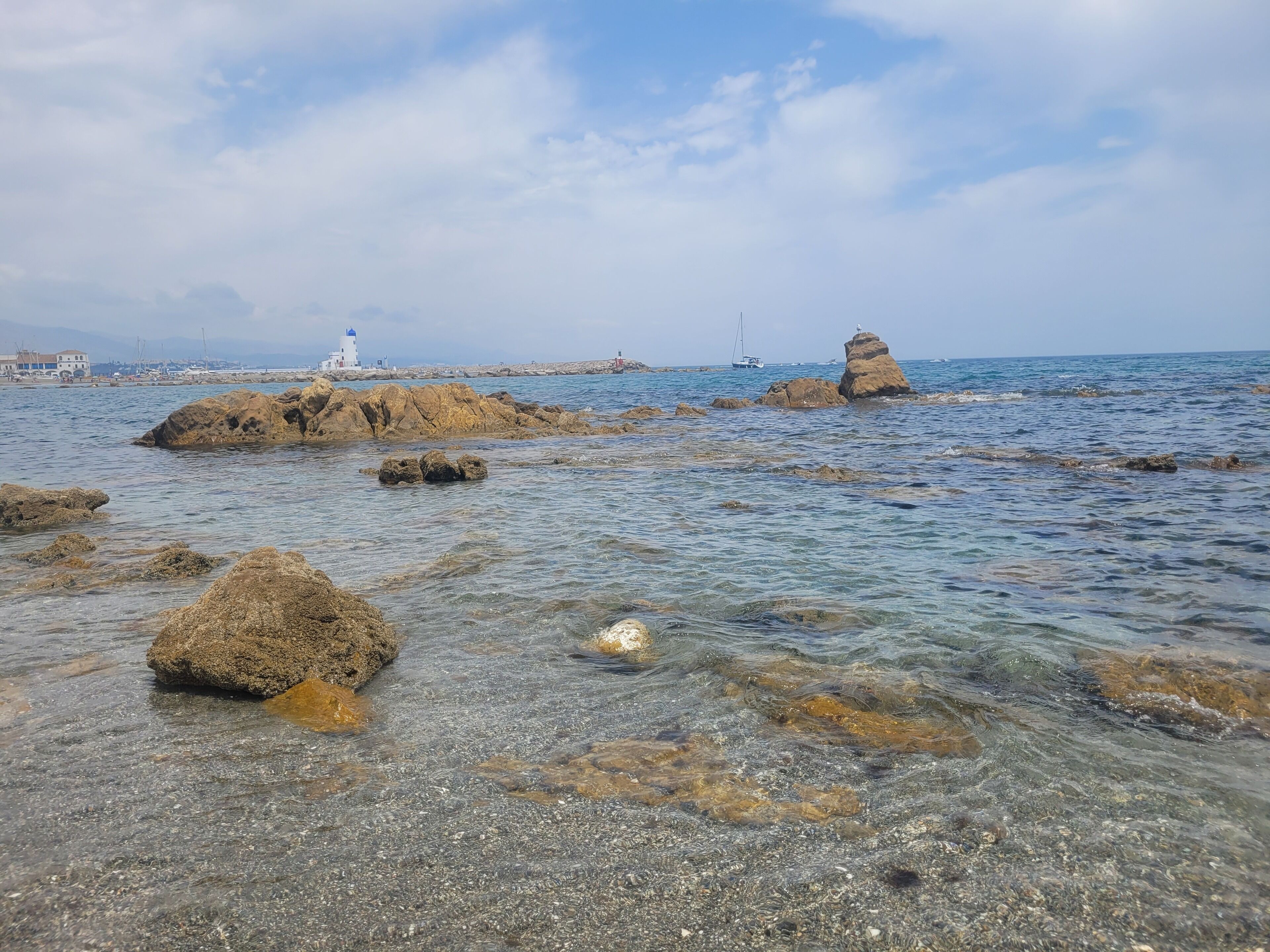 A rocky shoreline with clear water, a lighthouse, and a sailboat in the distance.