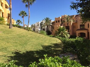 Eine angelegte Grünfläche mit einer grasbewachsenen Böschung, Palmen und Gebäuden im mediterranen Stil.