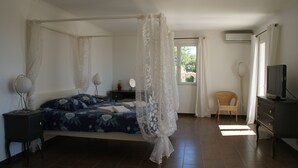A bedroom with a four-poster bed, a bedside table with a lamp, a chair, a television, and a window with curtains.