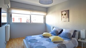 Un dormitorio con cama, almohadas, mesita de noche, lámpara, ventana y un cuadro colgado en la pared.