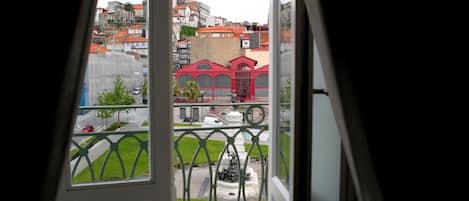 A view from a window with a metal railing, overlooking a street with buildings and a statue.