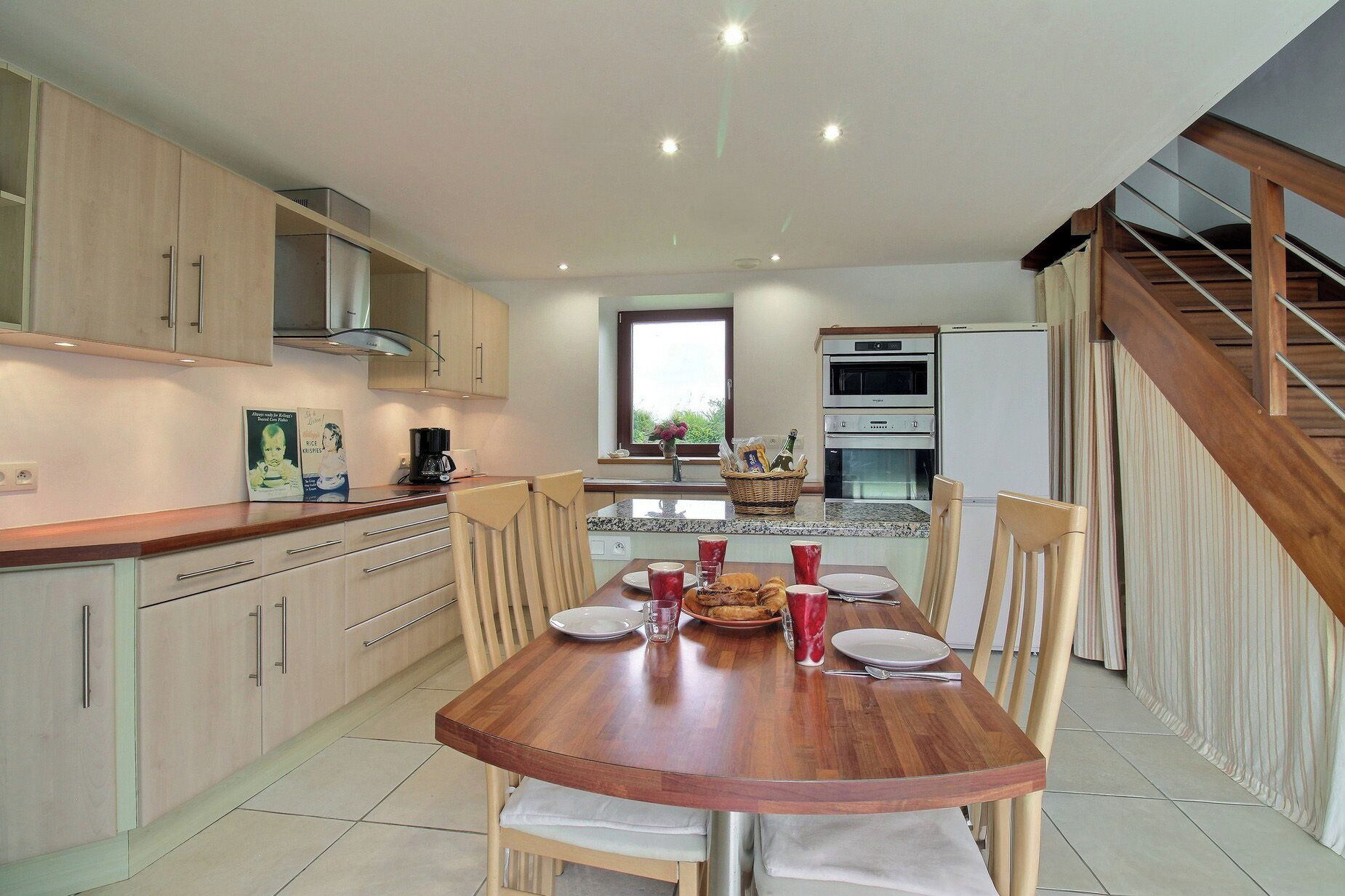 A modern kitchen with wooden cabinets, a dining table set for four, and a staircase leading to an upper level.