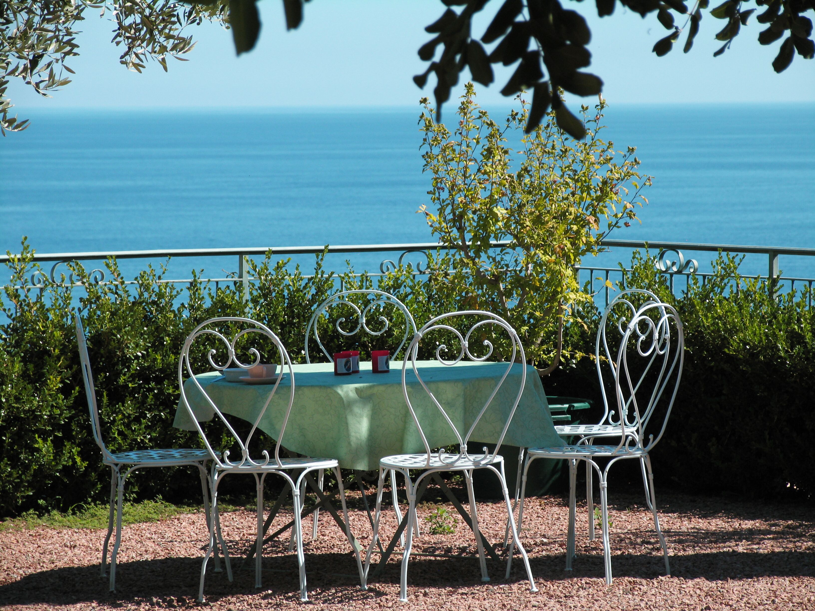 Een tafel met een groene tafelkleed en witte metalen stoelen, op een grindpad met uitzicht op zee en begroeiing op de achtergrond.
