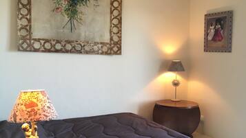 Een slaapkamer met een donker bed, een houten nachtkastje met een lamp, een lijst met bloemen en een kleiner fotolijstje aan de muur.