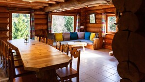 A log cabin interior with a wooden dining table, chairs, a sofa with colorful cushions, and large windows with curtains. - Superbe chalet rondins de 150 m² au cœur des Vosges, sauna, wifi, groupe, famille (La Bresse)