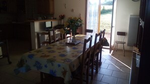 Un coin repas avec une table recouverte d’une nappe à motifs floraux, entourée de chaises en bois. On aperçoit un vase rempli de fleurs sur la table, ainsi qu’un écran d’ordinateur.