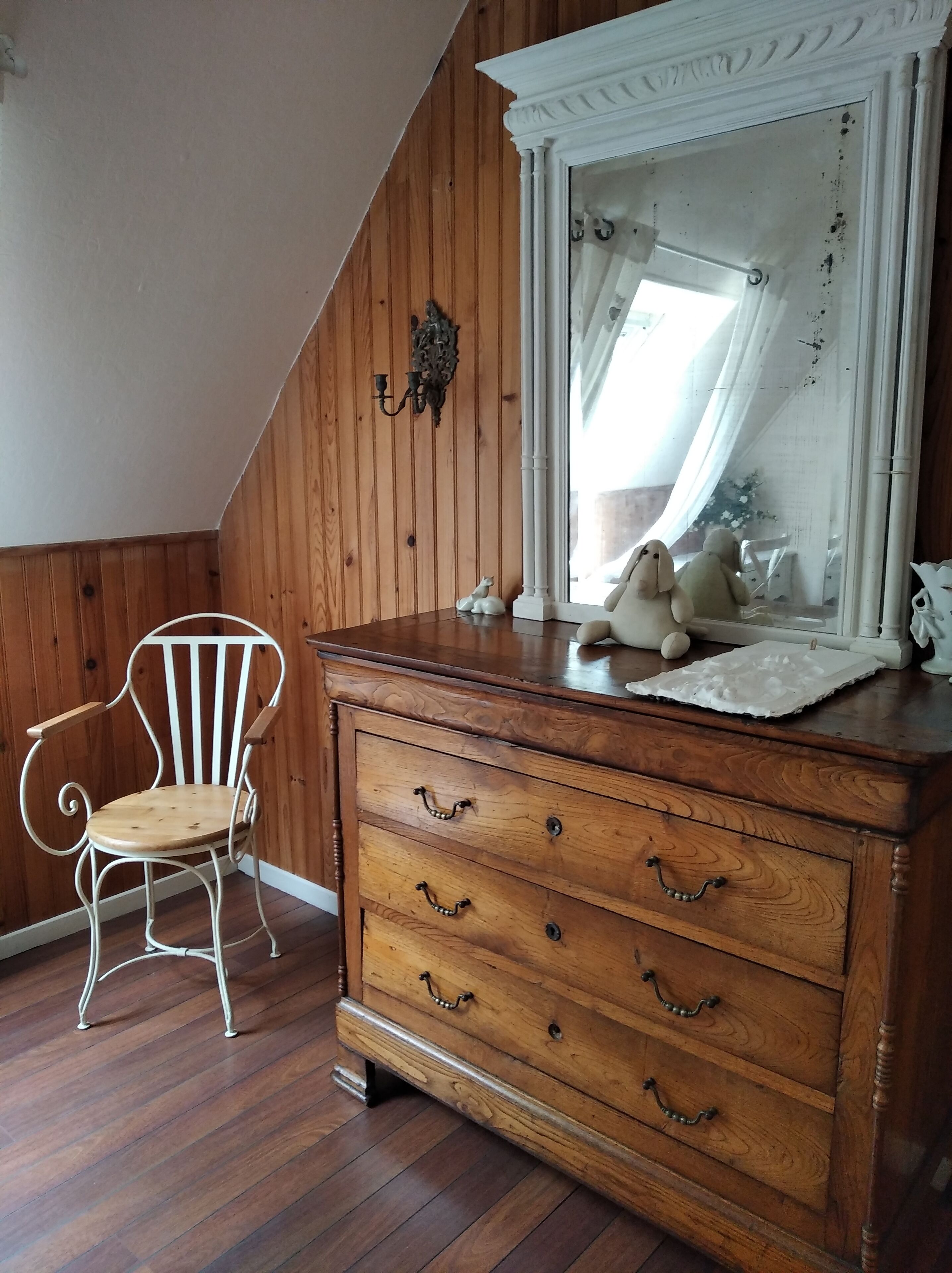 Une pièce aux murs lambrissés, avec une chaise blanche, une commode en bois dotée de tiroirs, un miroir et une petite statue blanche posée sur la commode.