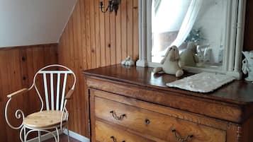 Une pièce aux murs lambrissés, avec une chaise blanche, une commode en bois dotée de tiroirs, un miroir et une petite statue blanche posée sur la commode.