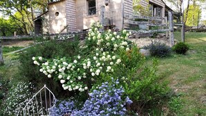 Un bâtiment rustique en pierre, doté d’un toit en pente, entouré d’une végétation luxuriante et de plantes en fleurs.