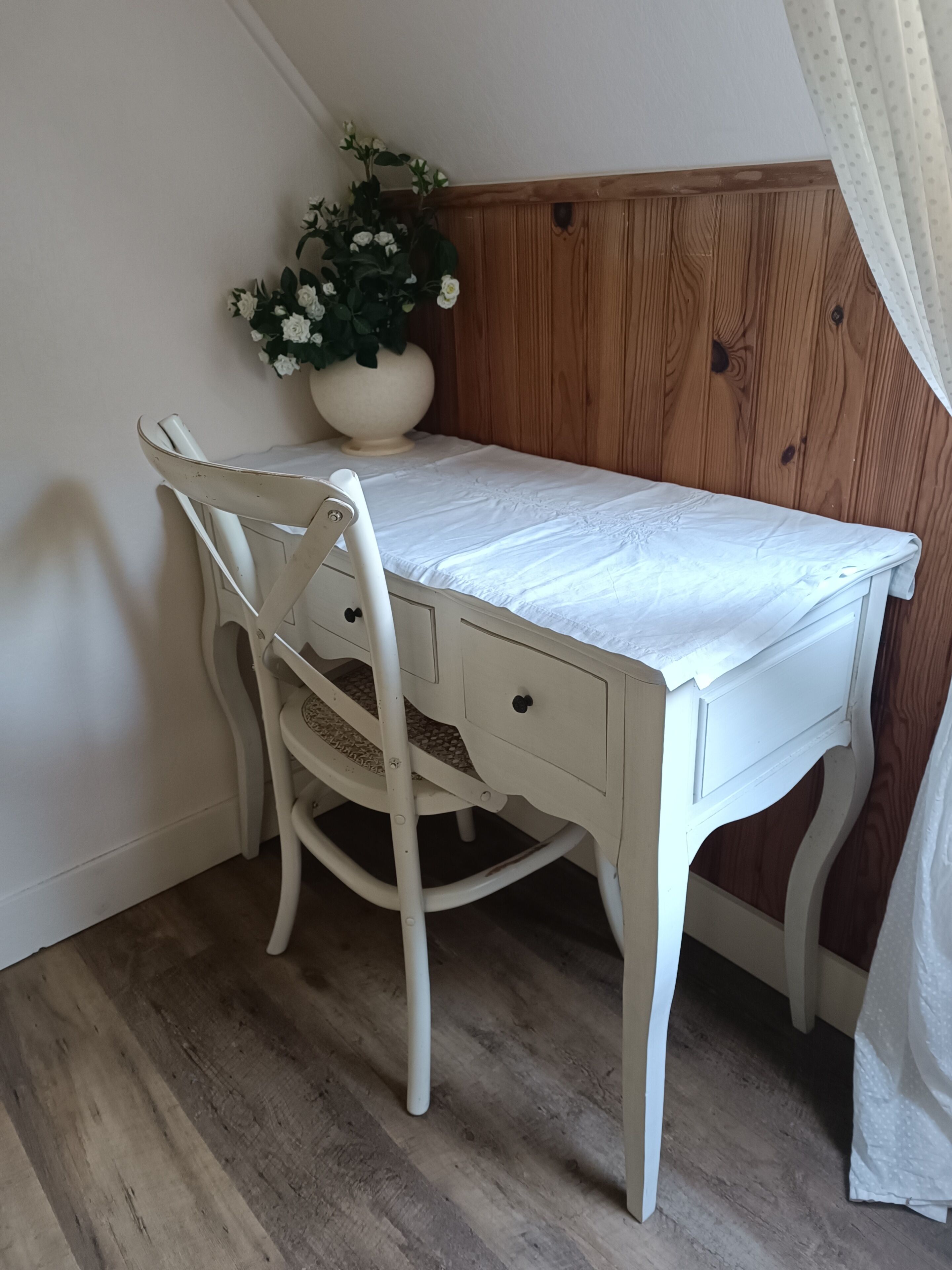 A white desk with a chair, a wooden panel wall, and a potted plant with white flowers.