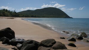 A beach with large rocks in the foreground, a sandy shore, and a mountain in the background. - House (Deshaies)