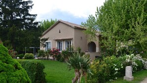 Une maison avec un toit en tuiles, un porche à l’avant et un jardin où poussent différentes plantes et fleurs.