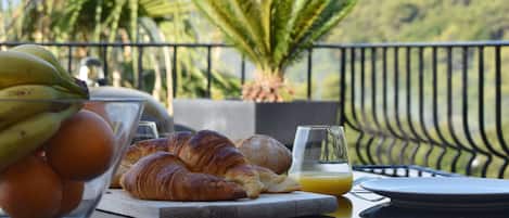 Desayuno con cruasanes, plátanos, naranjas y un vaso de zumo de naranja en un balcón con vistas a palmeras y montañas.