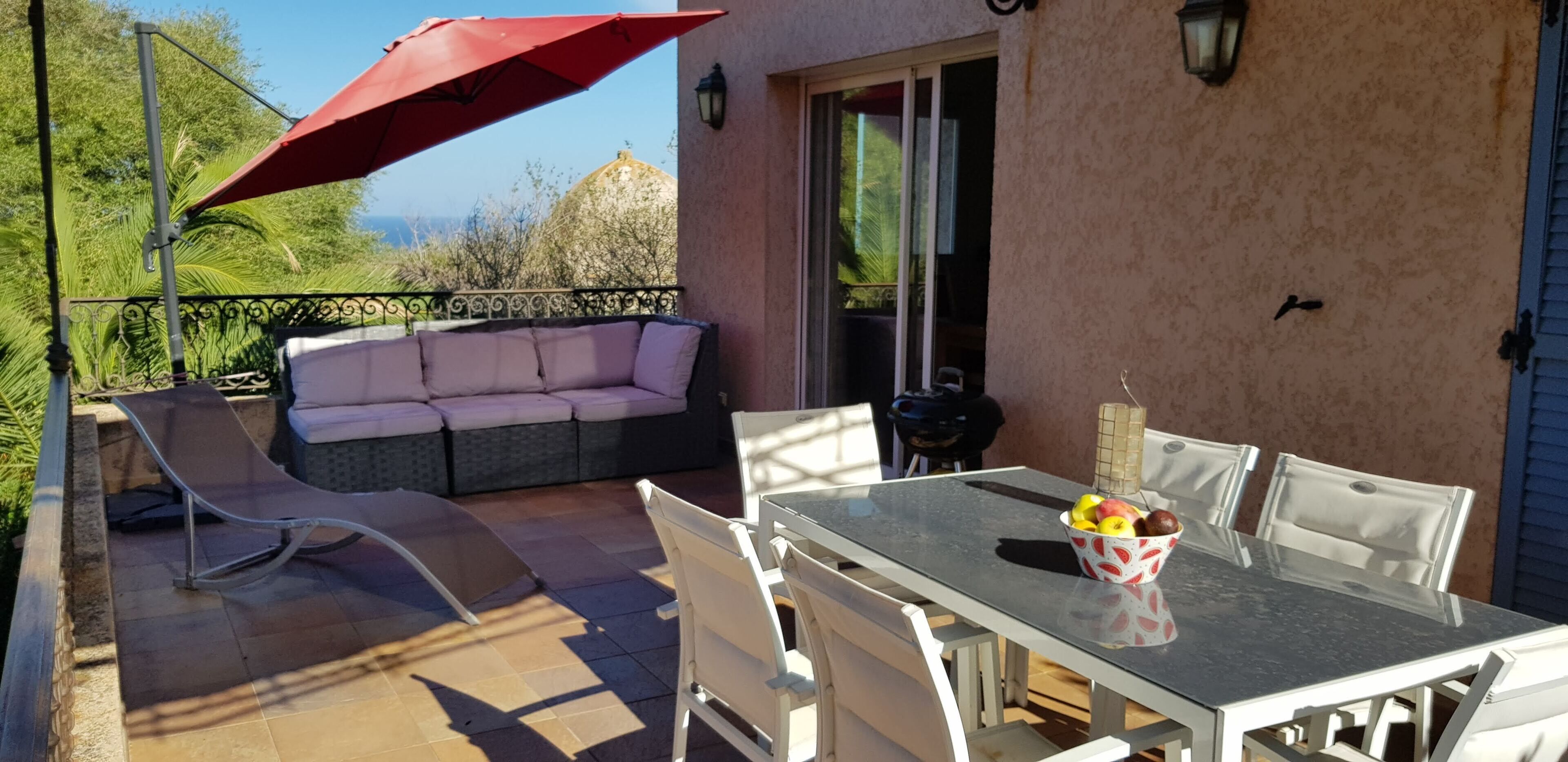 Patio esterno con tavolo in vetro, sedie bianche, un divano imbottito e un ombrello rosso.