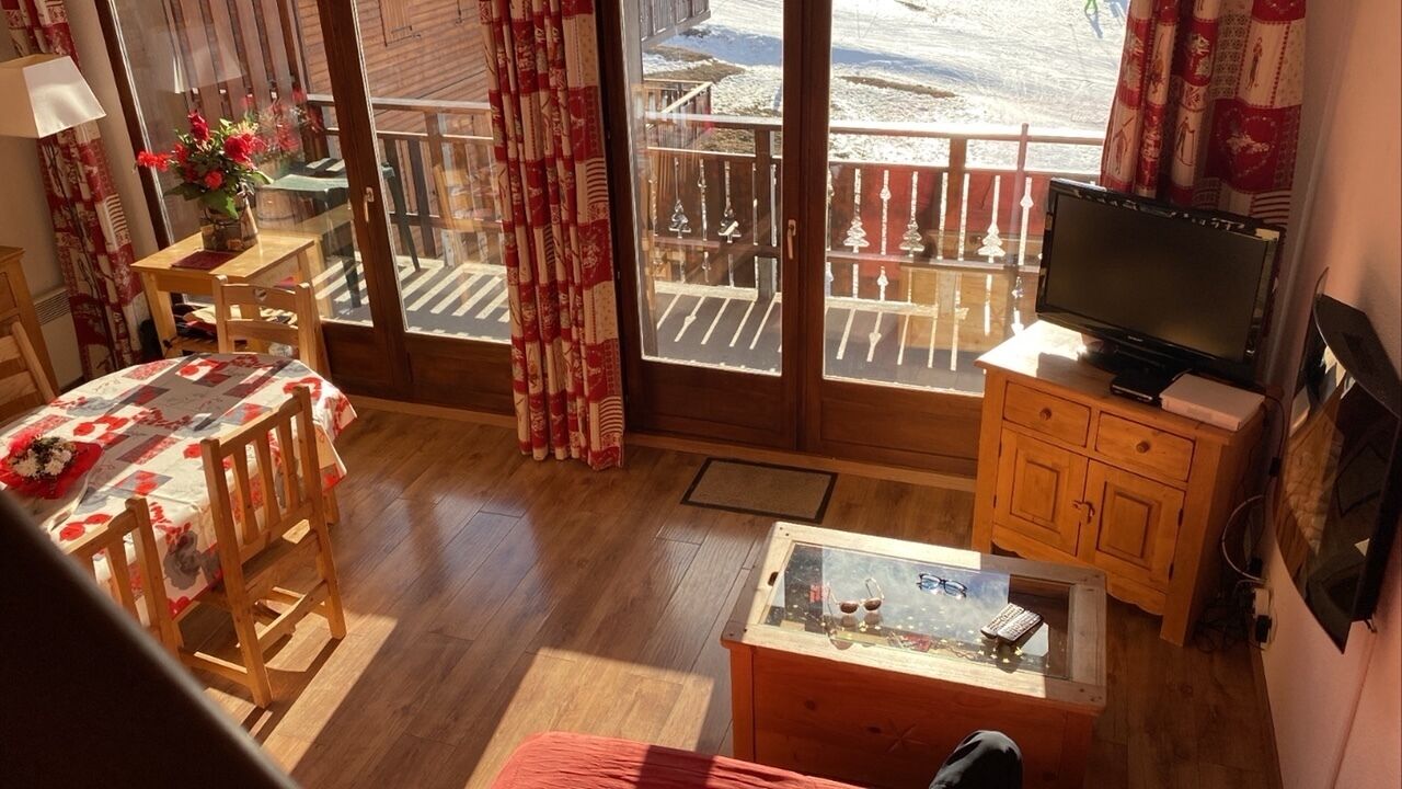 A cozy room with a wooden floor, a TV on a wooden cabinet, a table with chairs, a glass coffee table, and a balcony with a view of the outside.
