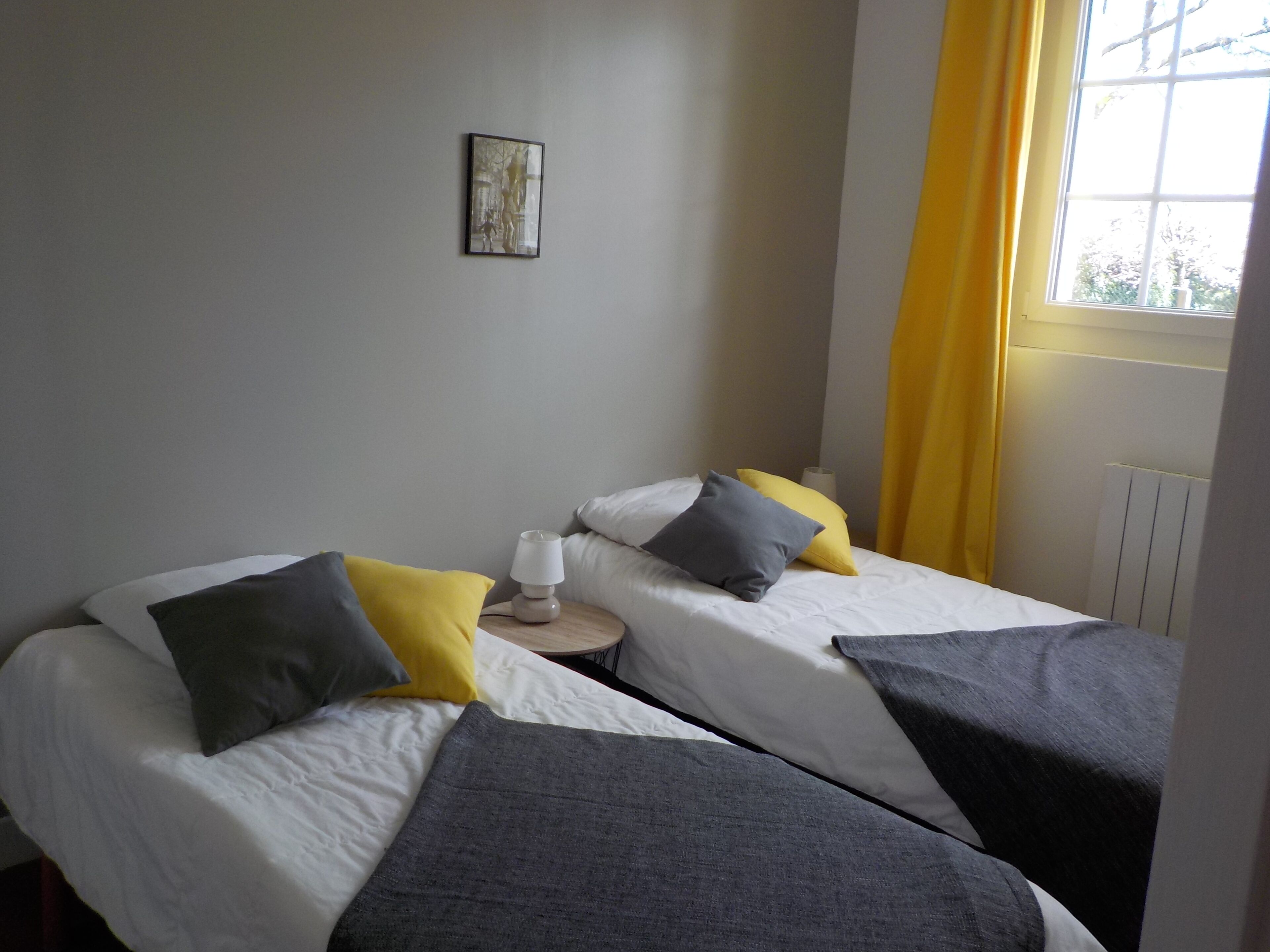 Una habitación con dos camas, cada una con una almohada y una mesita de noche con lámpara. Se ve una ventana con cortinas amarillas y un radiador en la pared.