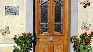 Une porte à deux battants en bois, dotée de panneaux de verre, flanquée de deux plantes en pot ornées de fleurs rouges, et un luminaire fixé au mur au-dessus.