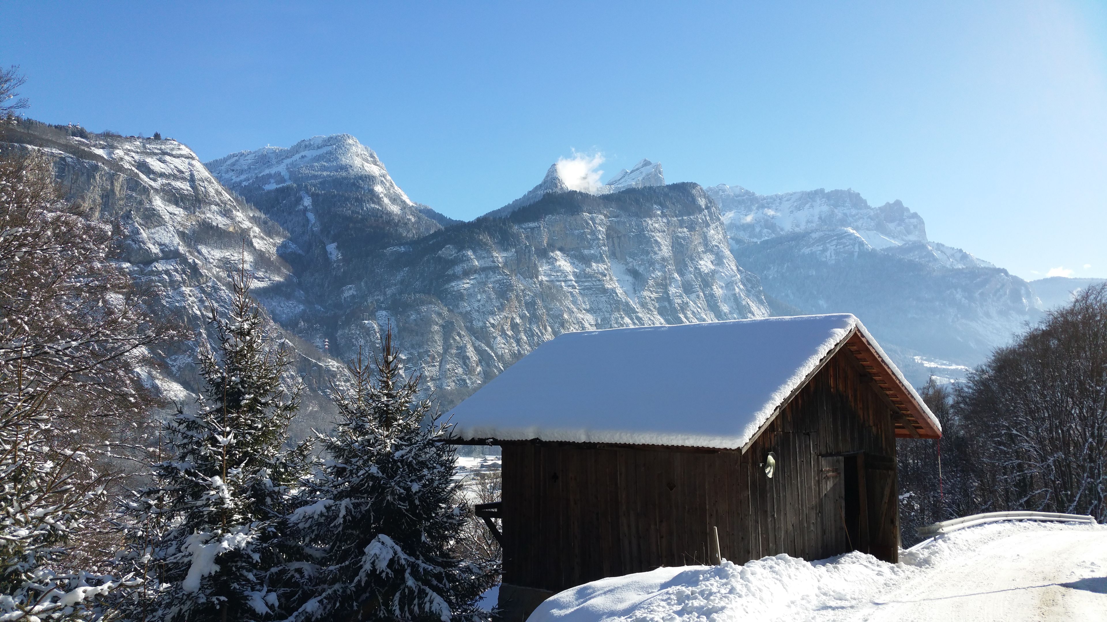 A wooden cabin with a snow-covered roof, surrounded by snow-laden trees and mountains in the background.