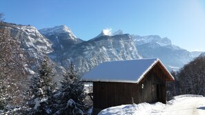 Una cabaña de madera con techo nevado, rodeada de árboles cargados de nieve y montañas al fondo.