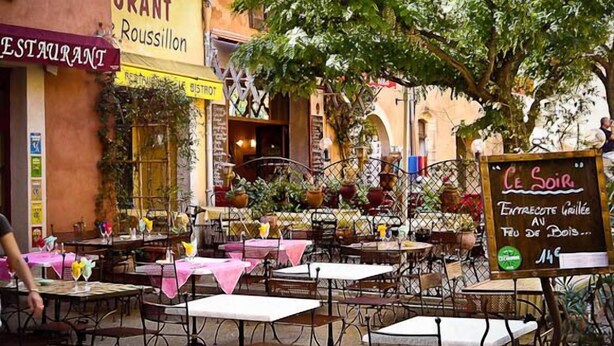 Un café en plein air avec des tables et des chaises, entouré de plantes et agrémenté d’une enseigne.