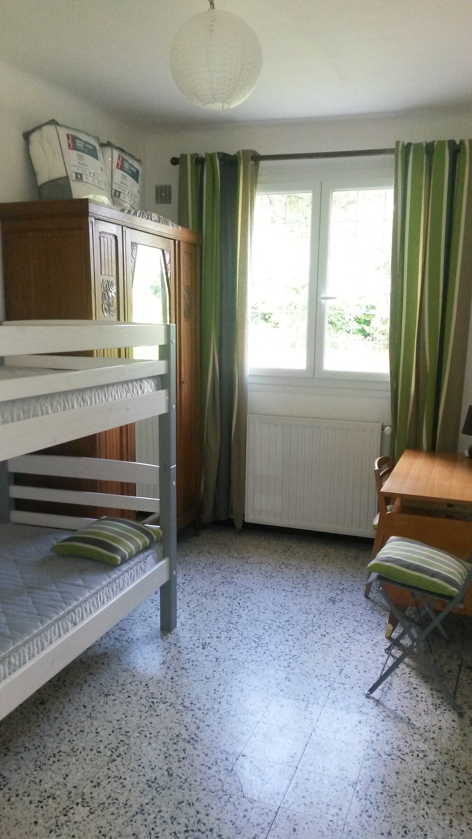 A room with bunk beds, a wooden cabinet, a radiator, a window with curtains, and a chair.