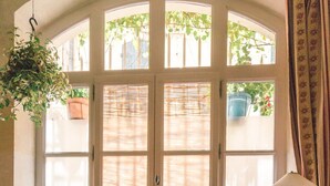 A room with a large arched window, a small table with a patterned tablecloth, a chair with a pink cushion, and a floor with a geometric tile pattern. - Charming studio 2 minutes from the Arena (Arles)